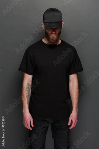 Hipster handsome male model with beard wearing black blank t-shirt and a black baseball cap with space for your logo or design in casual urban style