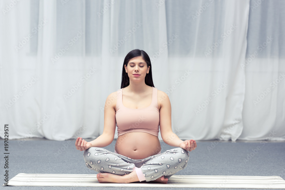 young pregnant woman with closed eyes sitting in lotus position on yoga mat