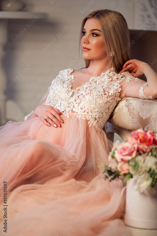 Naklejka premium Beautiful pregnant young woman is wearing luxury pink lingerie dress at home in cozy interior with bouquet of flowers