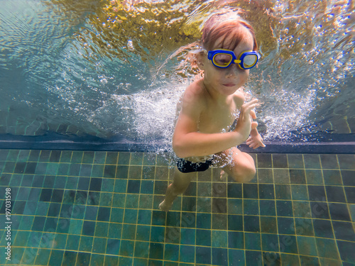 A child boy is swimming underwater in a pool, smiling and holding breath, with swimming glasses photo