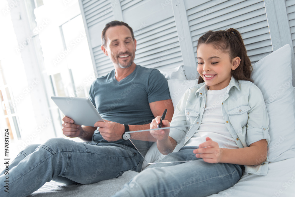 Smart child. Nice positive happy girl lying on the bed and using a tablet while spending time with her father Smart child. Nice positive happy girl lying on the bed and using a tablet while spending time with her father