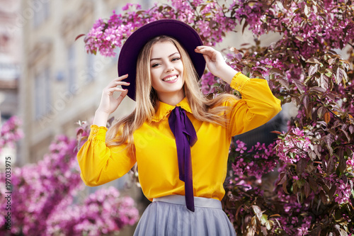 Outdoor portrait of young beautiful happy smiling girl posing in spring street with blooming pink trees. Model wearing stylish violet hat, bow tie, yellow blouse, skirt. Female fashion concept