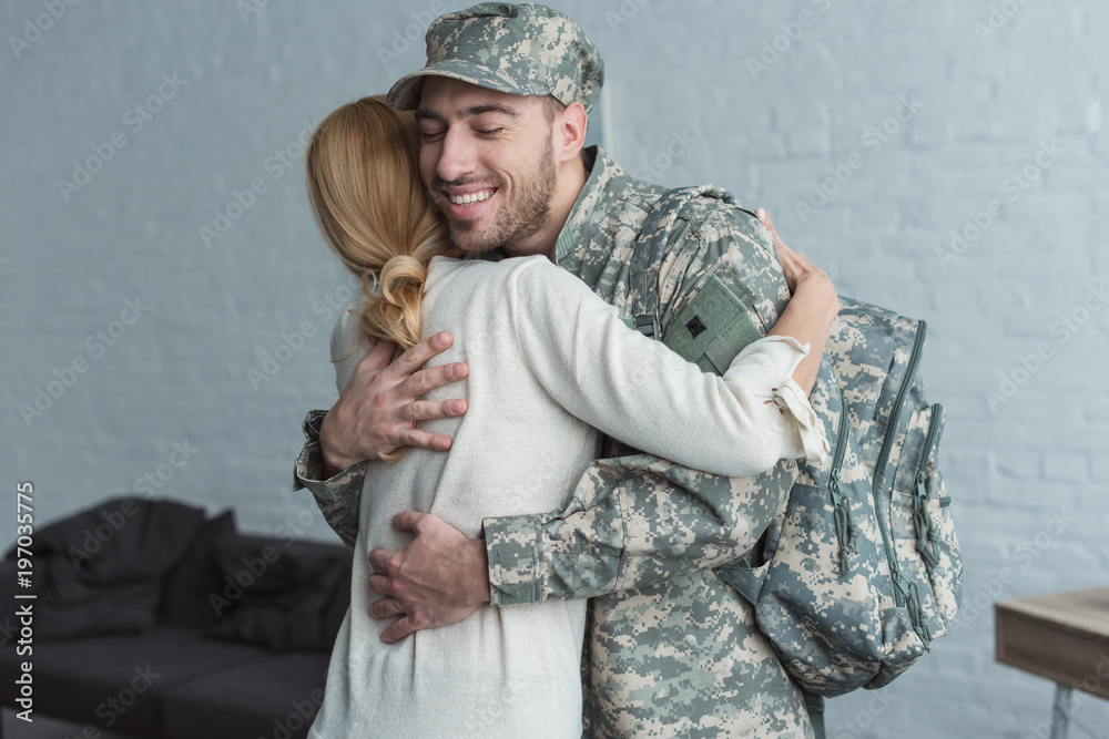 side view of smiling man in military uniform and mother hugging each other at home