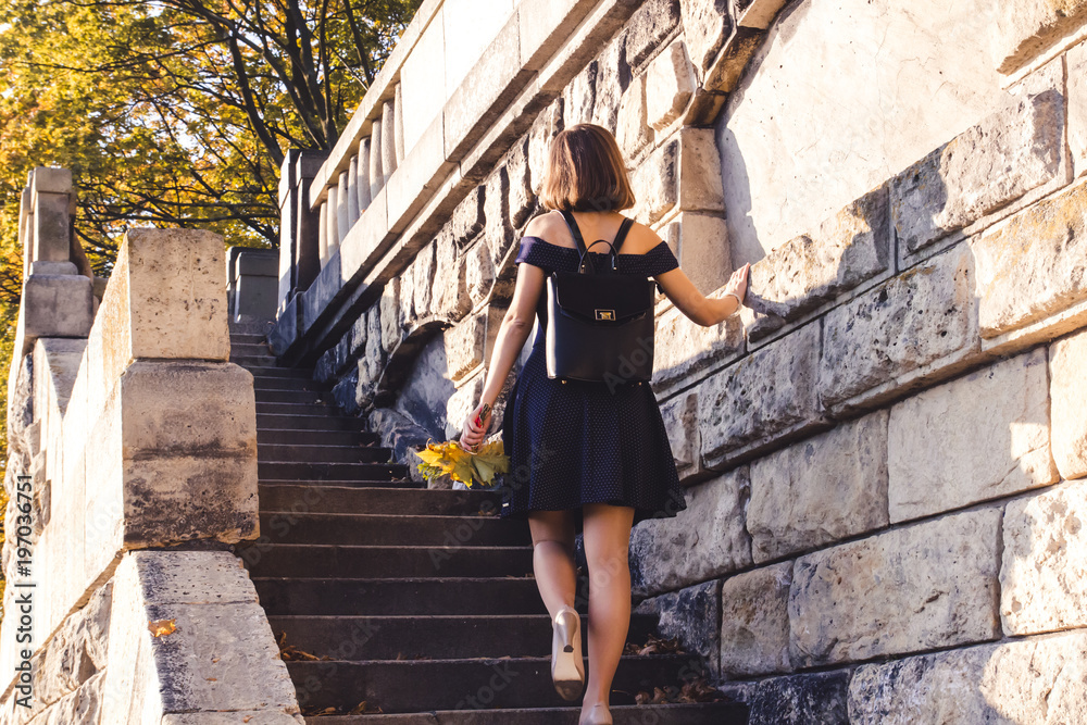 A young schoolgirl girl with a backpack and a bouquet of yellow autumn leaves walks down a large stone staircase to school