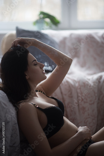 Sexy young woman in a black lingerie vaping in bedroom. Beautiful brunette woman smoking
