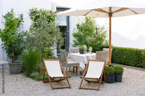Gravel terrace style townhouse, with wooden deckchair, umbrella in white fabric, table covered with linen and plants in pots photo