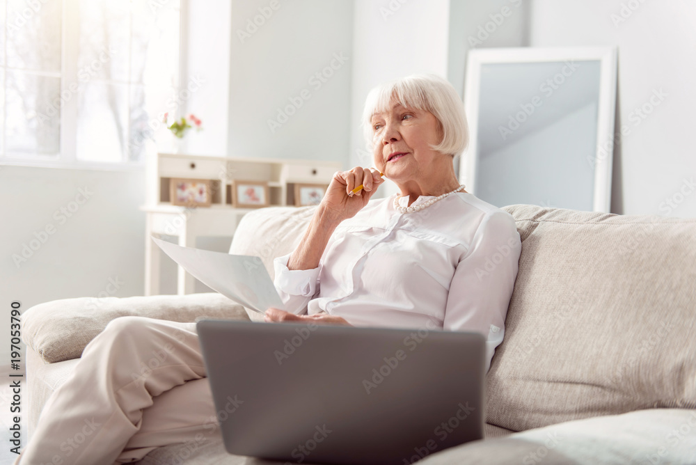 In search of ideas. Petite elderly woman sitting on the couch and being deep in thought, inventing the ways of improving her project In search of ideas. Petite elderly woman sitting on the couch and being deep in thought, inventing the ways of improving her project