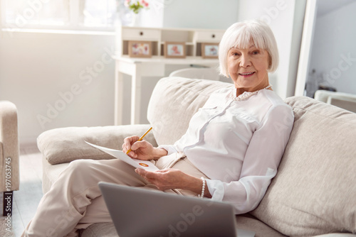 Skilled professional. Upbeat senior woman sitting on the sofa and posing for the camera while working on her business project and carrying out a market research © zinkevych
