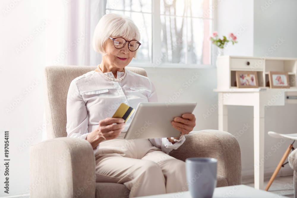 Enjoying convenience. Charming elderly lady sitting in the armchair in the living room and paying with her bank card for an online purchase Enjoying convenience. Charming elderly lady sitting in the armchair in the living room and paying with her bank card for an online purchase