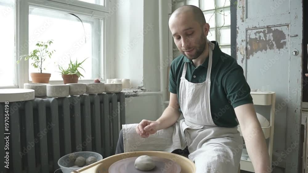 Portrait of male ceramist, who is starting his work in bright studio. Man in dickey and wirh beard is sitting at throwing wheel, putting piece of clay to it, turning on the equipment, pouring watter