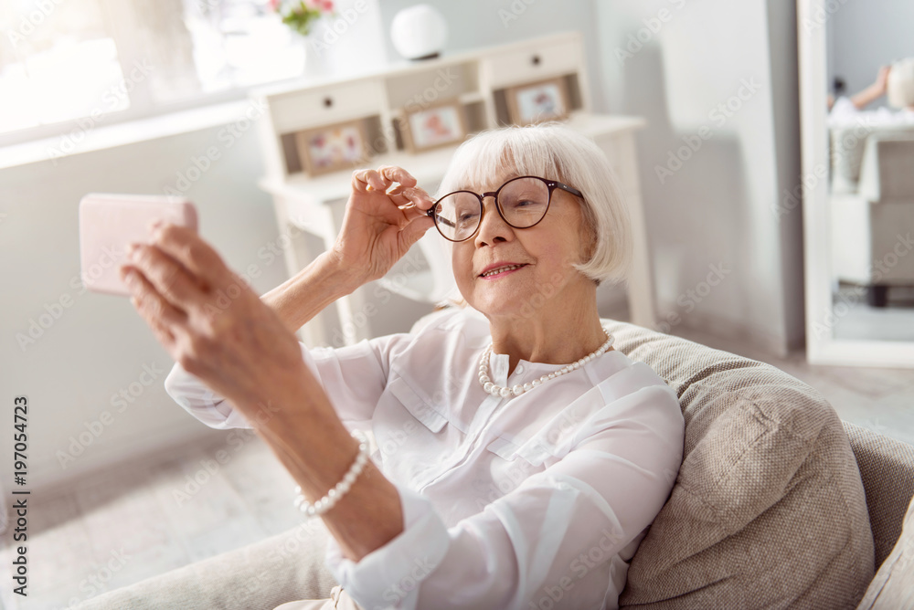 Obraz premium Perfect mood. Cheerful senior woman sitting on the sofa and smiling at the camera, adjusting her eyeglasses, while taking selfie in her living room