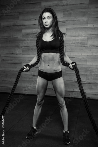 Stunning fit girl with black hair in the black short top, shorts and gloves training with rope in a gym, wooden wall at background. Black and white portrait.