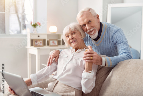 Pleasant distraction. Joyful elderly man hugging his beloved wife from behind while she sitting on the couch and working on the laptop