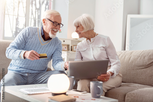 We are shopaholics. Upbeat elderly couple shopping online together and the man pointing at the laptop, choosing the item to buy, while giving his bank card to pay for it