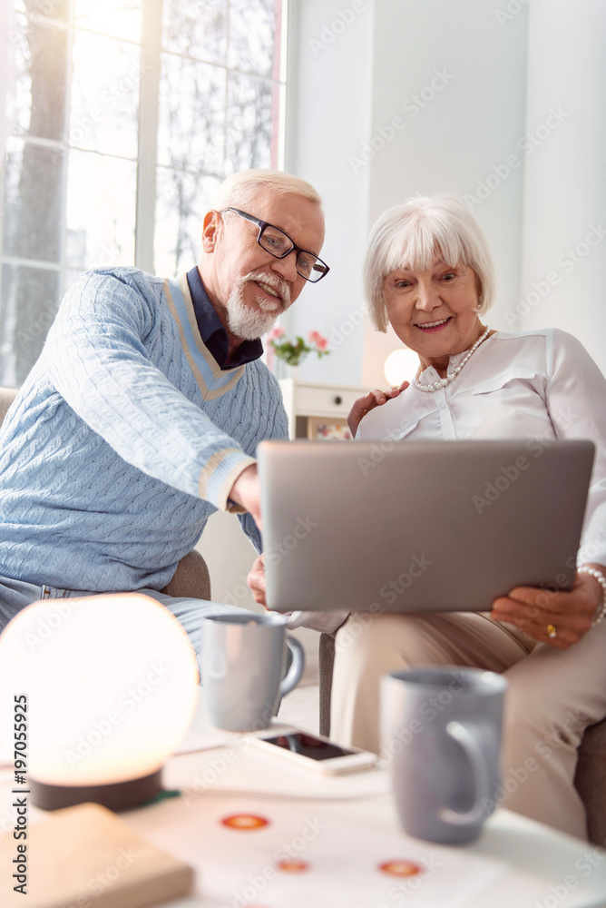 Having fun. Upbeat senior couple sitting in the living room and watching a funny video on their laptop while the man pointing at his favorite moment