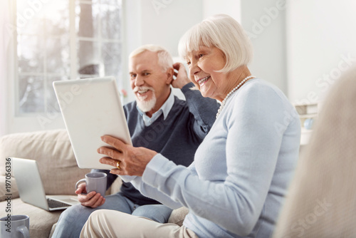 Quality time together. Upbeat elderly husband and wife sitting in the living room and watching a video on tablet together while smiling widely