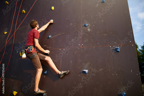 Sporty man practicing rock climbing in gym on artificial rock training wall outdoors. Young talanted slim climber guy on workout. © artiemedvedev