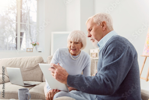 Share your opinion. Pleasant bearded elderly man showing his wife a tablet with a social media post open on it and discussing it together with her