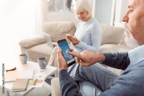 Useful app. The focus being on a bearded elderly man sitting in the armchair in the living room and checking the weather forecast in the application on his phone © zinkevych