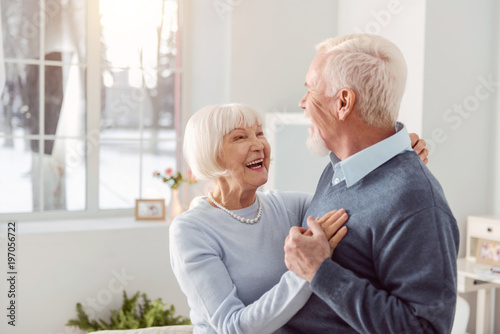 Loving couple. Joyful elderly husband and wife dancing in the living room while smiling at each other widely