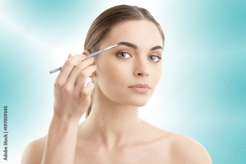 Studio shot of a beautiful young woman applying makeup to her eyebrow while standing at isolated background.