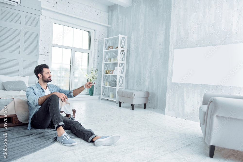 Here I am. Cheerful young man keeping smile on his face and stretching left arm while watching television