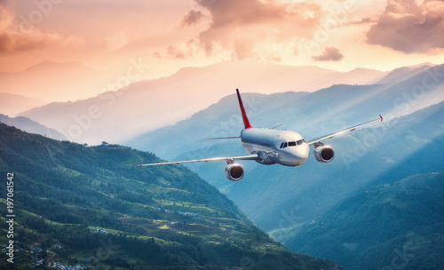 Aircraft is flying over green hills against mountains with yellow sunbeams at sunset. Landscape with passenger airplane, colorful sky, village. Passenger aircraft. Business travel. Commercial plane