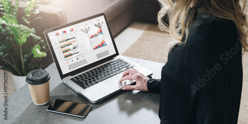 Rear view. Young businesswoman is sitting at table, working on laptop with graphs, charts, diagrams, schedules on screen