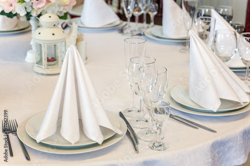 Glasses, flowers, fork, knife, napkin folded in a pyramid, served for dinner in restaurant with cozy interior. Wedding decorations and items for food, arranged by the catering service on a large table © Юлия Завалишина