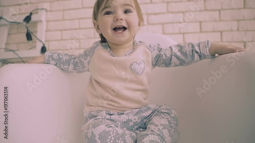 Cheerful little brunette baby girl sitting on white chair with smile slow motion. the child tries to reach out to the camera.