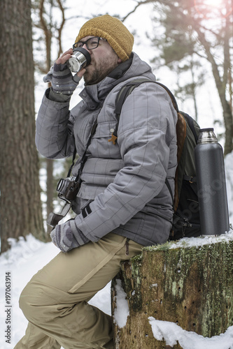 Handsome, bearded man in glasses with a camera and a backpack sitting on a tree stump in the winter forest and is holding a cup with a hot drink from a thermos.