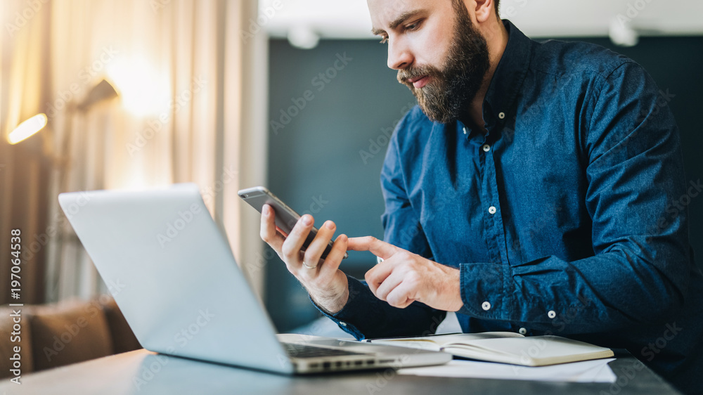 Smiling bearded businessman sitting at table in front of computer, using smartphone.Freelancer works home, distance work Smiling bearded businessman sitting at table in front of computer, using smartphone.Freelancer works home, distance work