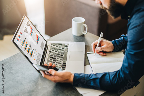 Bearded businessman sitting at table in front of laptop with graphs,charts,diagrams on screen, making notes in notebook.