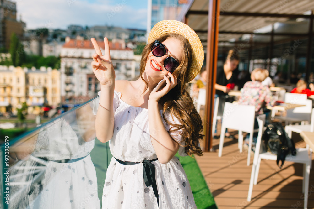 Fototapeta premium Cute girl with long hair in sunglasses is standing on the terrace. She wears a white dress with bare shoulders, red lipstick and hat . She is speaking on phone and smiling to the camera.