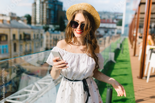 Cute girl with long hair  in sunglasses is standing on the terrace. She wears a white dress with bare shoulders  red lipstick  and hat . She is hearing music through headphones.
