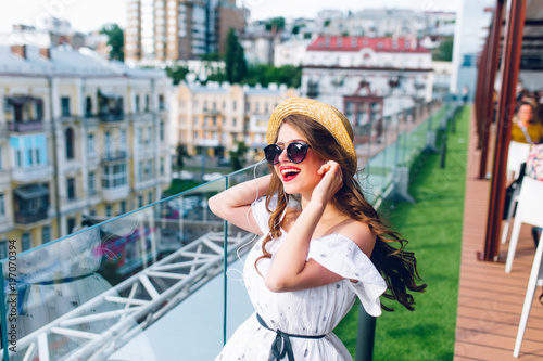 Fototapeta Naklejka Na Ścianę i Meble -  Happy girl with long hair  in sunglasses is listening to music through headphones on the terrace. She wears a white dress with bare shoulders, red lipstick  and hat . 