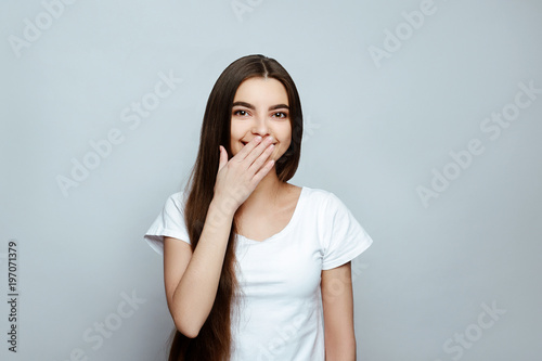 human emotions. Portrait of the young beautiful woman brunette with long dark brilliant hair. The girl poses on a white background, standing with folded arms and smiling have closed a mouth a hand