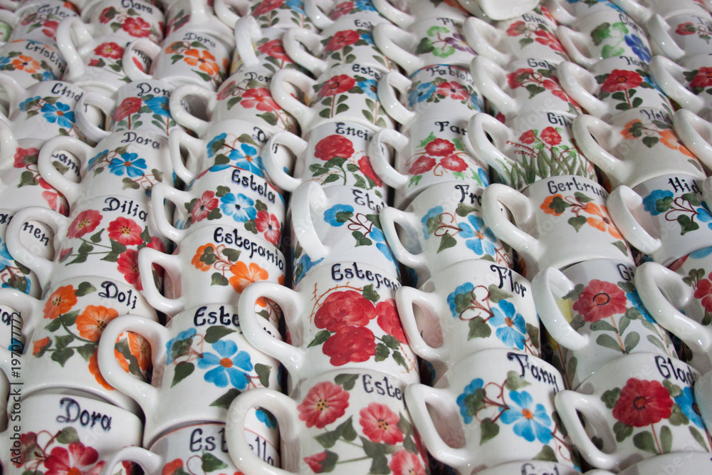 Bunch of ceramic mugs with names. Stacked ceramic mugs. Mugs with women names and colorful flowers handcrafted. Industrial, Production, and ManuFacturing Concept. Bunch of ceramic mugs with names. Stacked ceramic mugs. Mugs with women names and colorful flowers handcrafted. Industrial, Production, and ManuFacturing Concept.