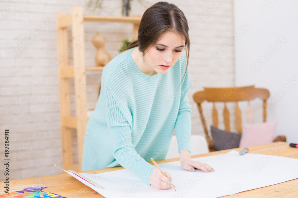 Attractive young architect in casualwear standing at wooden desk and working on blueprint, interior of modern office on background