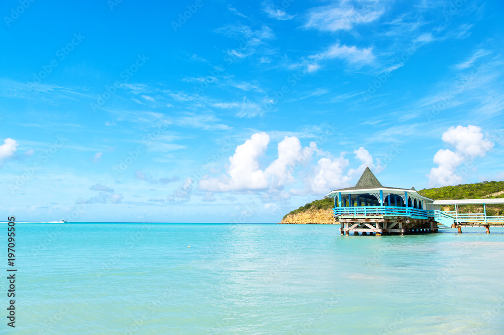 Obraz premium Sea beach with wooden shelter on sunny day in antigua. Pier in turquoise water on blue sky background. Summer vacation on caribbean. Wanderlust, travel, trip. Adventure, discovery, journey