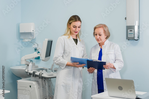 Senior doctor talking with young woman assistant standing in the gynecological office with chair and lamp on the background