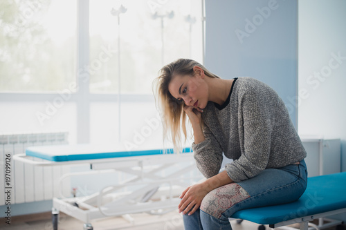 Young pregnant woman sitting on bed in comfortable ward, waiting for doctor thoughtfully