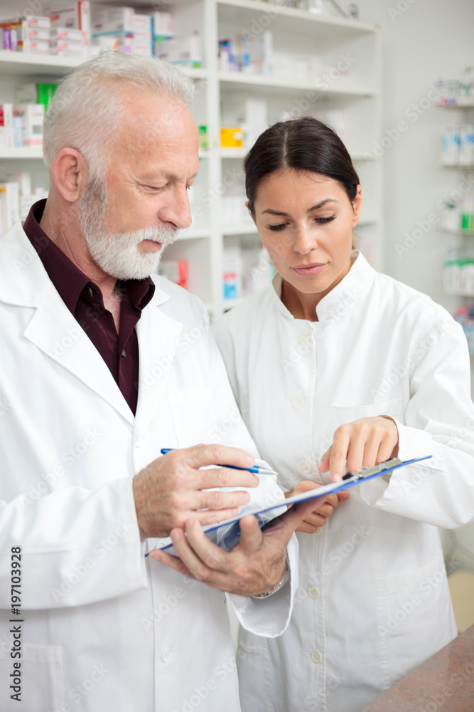 Medicine, pharmaceutics, health care and people concept - Serious young female and senior male pharmacists writing to a clipboard.