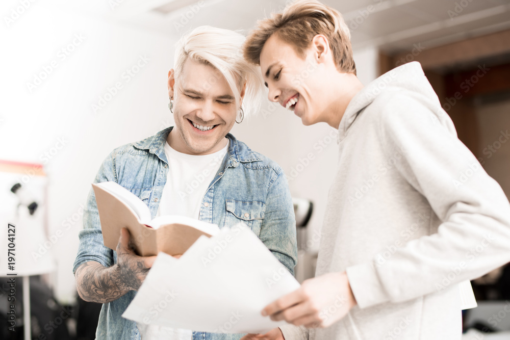 Obraz premium Caucasian hipster-like tattooed male student and his friend looking at pictures in textbook and smiling joyfully