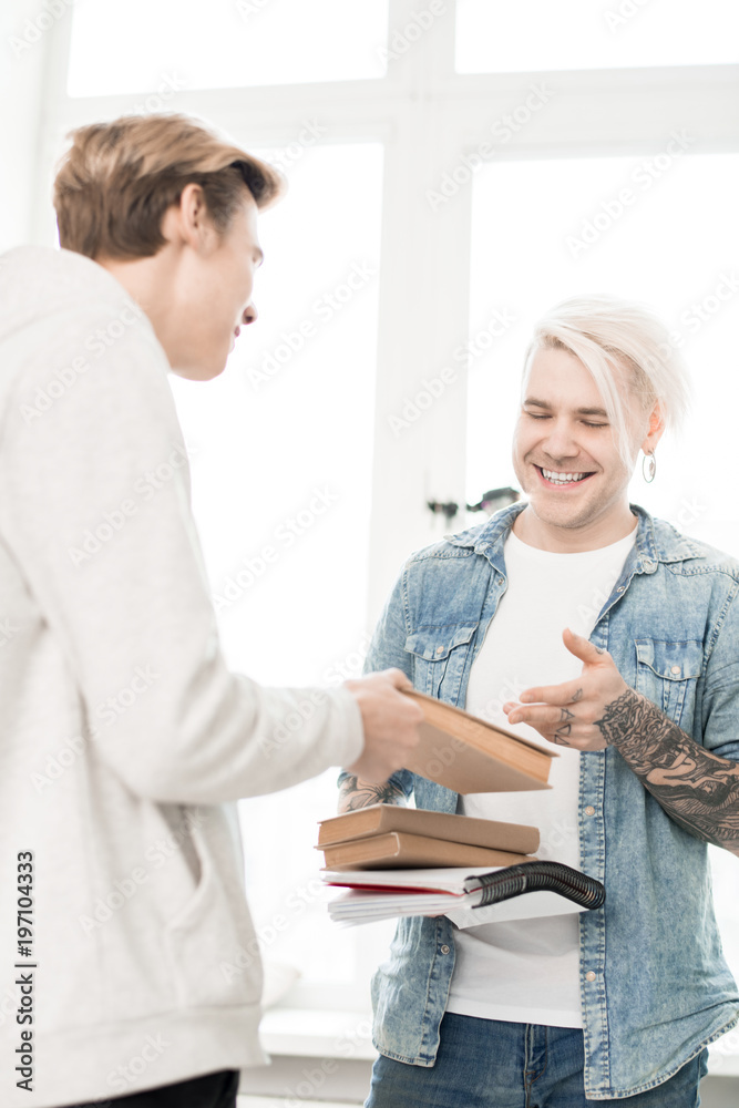 Obraz premium Tattooed adult student with dyed blond hair exchanging books with his young college mate and smiling happily, low angle view