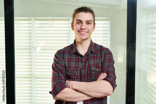Waist-up portrait of young Caucasian man in casual shirt standing in office with his hands crossed and smiling at camera happily