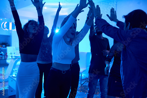 Group of modern young people dancing listening to DJ playing music at private house party, focus on beautiful girl high five with DJ lit by blue light