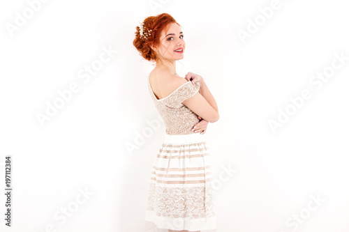 young girl with red hair in a beige lace dress standing from the back and smiling on a white wall background
