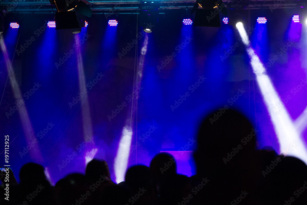 Concert crowd attending a concert, people silhouettes are visible, backlit by stage blue lights. Smart phones are visible here and there.