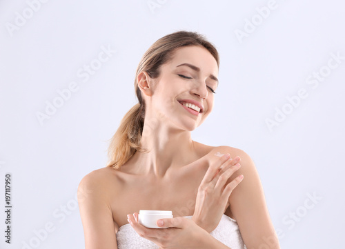 Young woman applying body cream on light background